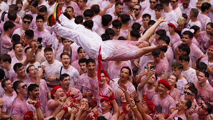 Multitud en Sanfermines 2025 elevando a un hombre; todos vestidos de blanco y rojo, manchados de vino, ambiente festivo.