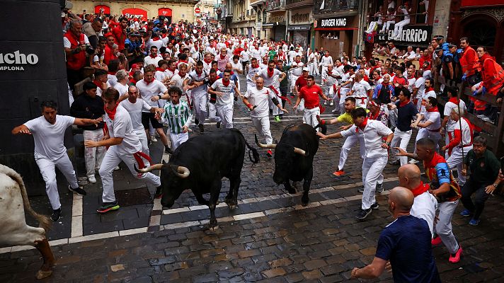 Multitud corriendo por una calle adoquinada durante un encierro, esquivando dos toros negros.  Espectadores en balcones y aceras.