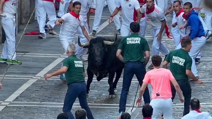 Toro negro en encierro, corredores blancos con pañuelos rojos, pastores con camisetas verdes \"PASTORES UNAIAK\" guiando. Calle adoquinada.