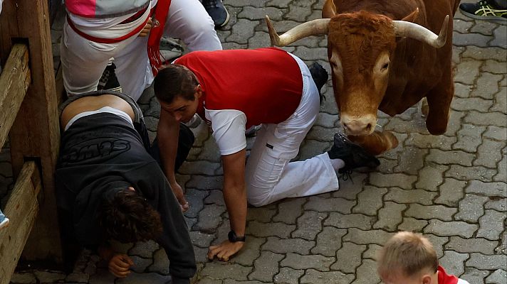 Toro marrón claro embiste a corredores durante un encierro. Uno está en el suelo, con camiseta roja y pantalones blancos, buscando refugio junto a otro corredor y una valla.