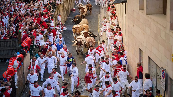 Multitud corriendo por una calle estrecha perseguida por toros.  Algunos corredores miran hacia atrás. Se observa personal de seguridad cerca.