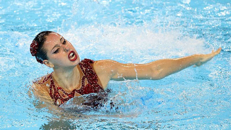 Nadadora de natación artística en acción; traje granate, pelo recogido, expresión concentrada; agua azul clara con salpicaduras.