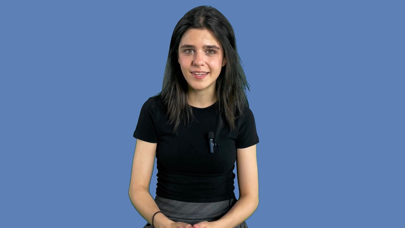 Mujer con micrófono de solapa, sentada frente a cámara con fondo azul.  Cabello oscuro y largo, viste camiseta negra y falda gris.