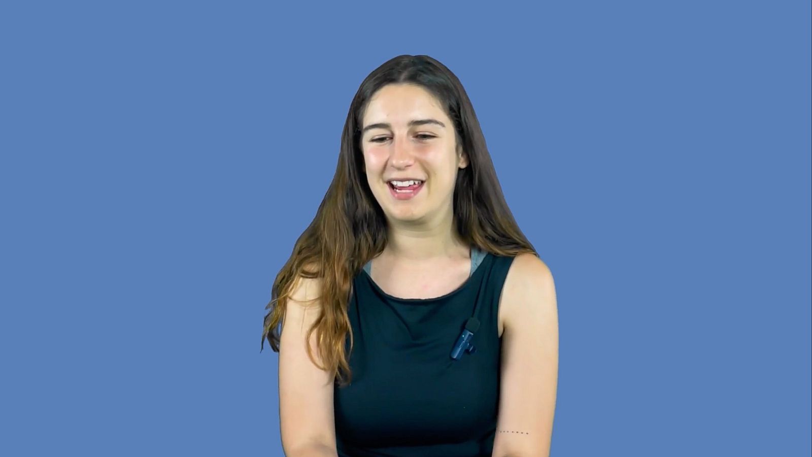 Mujer con camiseta negra y micrófono de solapa, sonriendo y mirando hacia abajo a la izquierda, en un fondo azul.