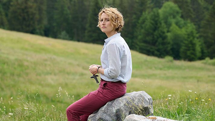 Mujer sentada en roca, prado y bosque al fondo.  Vista previa de película o programa, ambiente tranquilo y natural.