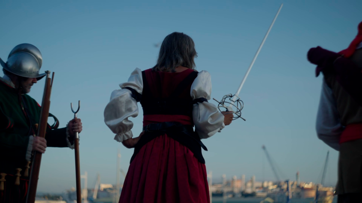 Una escena con tres personajes vestidos con trajes de época, uno con armadura y mosquete, otro con vestido rojo y negro empuñando una espada, y un tercero con vestimenta similar. El fondo muestra una ciudad portuaria.