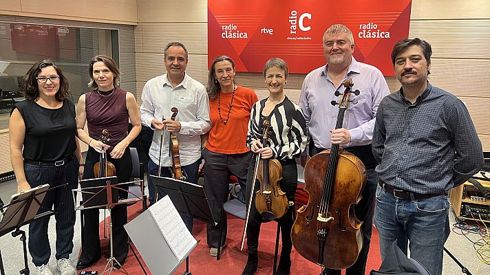 En un estudio de radio, seis personas posan para una fotografía. Se observan atriles con partituras y varios instrumentos musicales, como violines y un violonchelo, sugiriendo una sesión de grabación o entrevista.