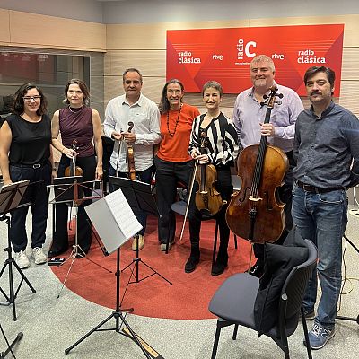 En un estudio de radio, seis personas posan para una fotografía. Se observan atriles con partituras y varios instrumentos musicales, como violines y un violonchelo, sugiriendo una sesión de grabación o entrevista.