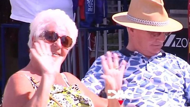 Una pareja de ancianos sonríe y saluda. La mujer lleva gafas de sol y un vestido floral, mientras que el hombre lleva un sombrero y una camisa estampada. Se observa una estructura metálica y una bolsa con la palabra "Sun".