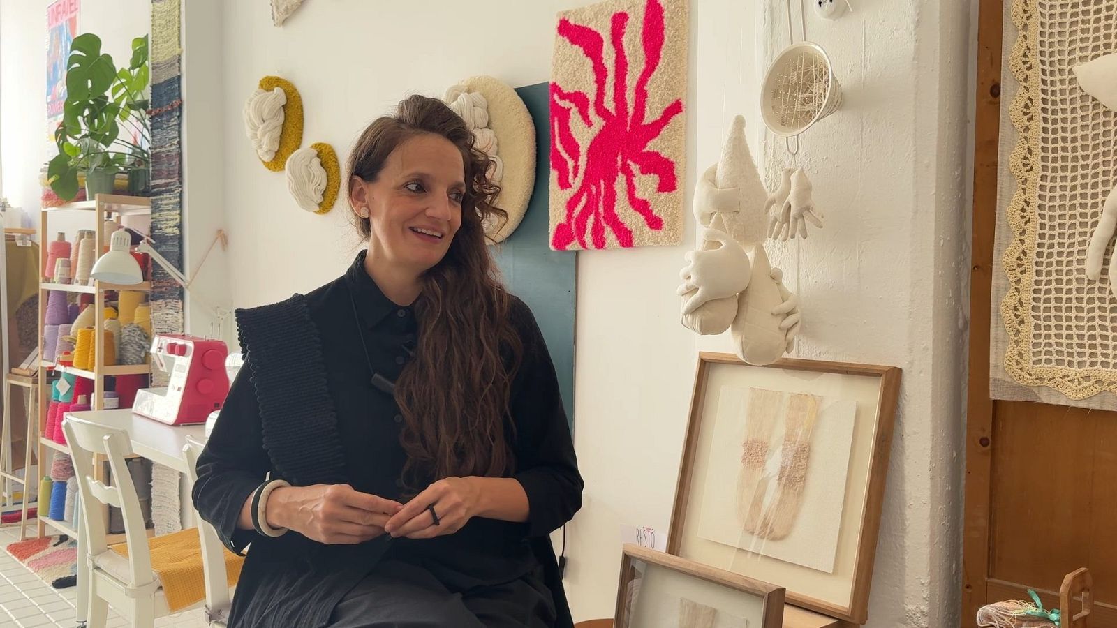 Una mujer sonriente con cabello castaño y blusa negra posa frente a un fondo decorado con obras de arte y elementos relacionados con la costura, como textiles y una máquina de coser.