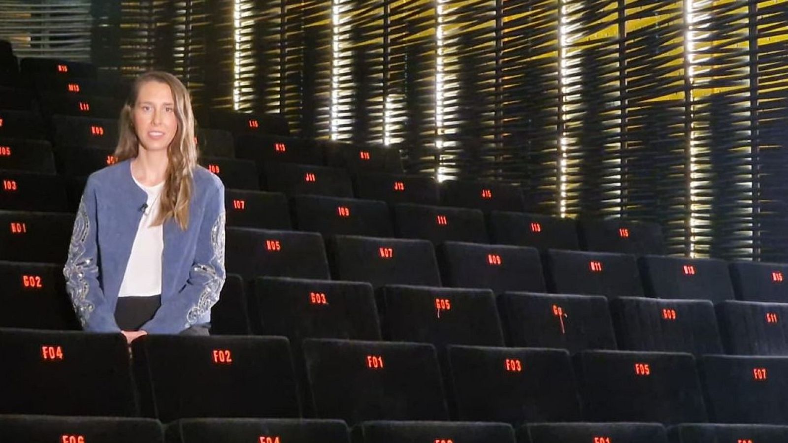 En una sala de cine vacía, una mujer con chaqueta azul y cabello castaño observa la pantalla. Las butacas negras están numeradas en rojo, y el fondo presenta un diseño de listones de madera iluminados.