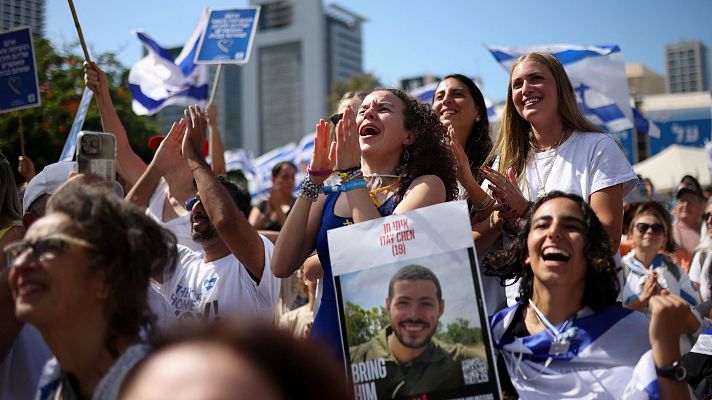 Una manifestación en una ciudad israelí muestra a una multitud ondeando banderas y carteles, con personas expresando alegría y esperanza. Una mujer sostiene un cartel con la frase \"Bring Him Home\", posiblemente en apoyo a la liberación de rehenes.