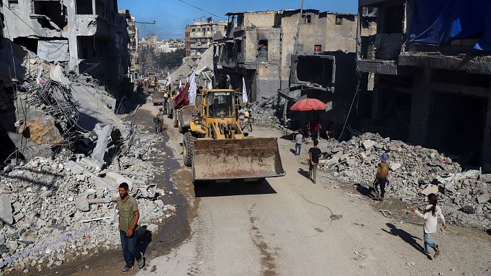 Una calle en Gaza, devastada por la destrucción, con edificios dañados y escombros. Una excavadora avanza, seguida por personas con banderas, mientras otras transitan por la zona afectada.
