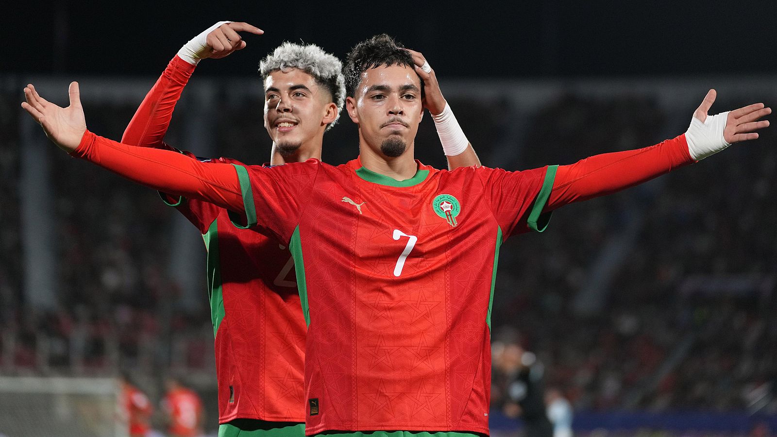 Dos futbolistas con camisetas rojas y detalles verdes celebran un gol, posiblemente en un partido de fútbol. Uno de ellos lleva el número 7 y el otro señala hacia arriba.