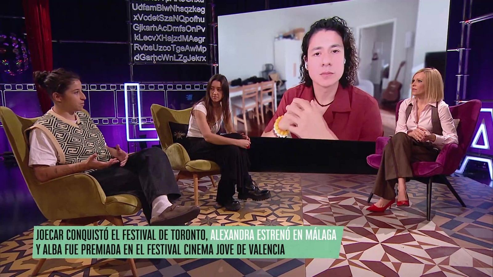 En un estudio televisivo, tres personas están sentadas en sillas de diferentes colores, mientras una pantalla muestra a un hombre en videollamada. El suelo presenta un diseño geométrico y un titular anuncia eventos relacionados con festivales.