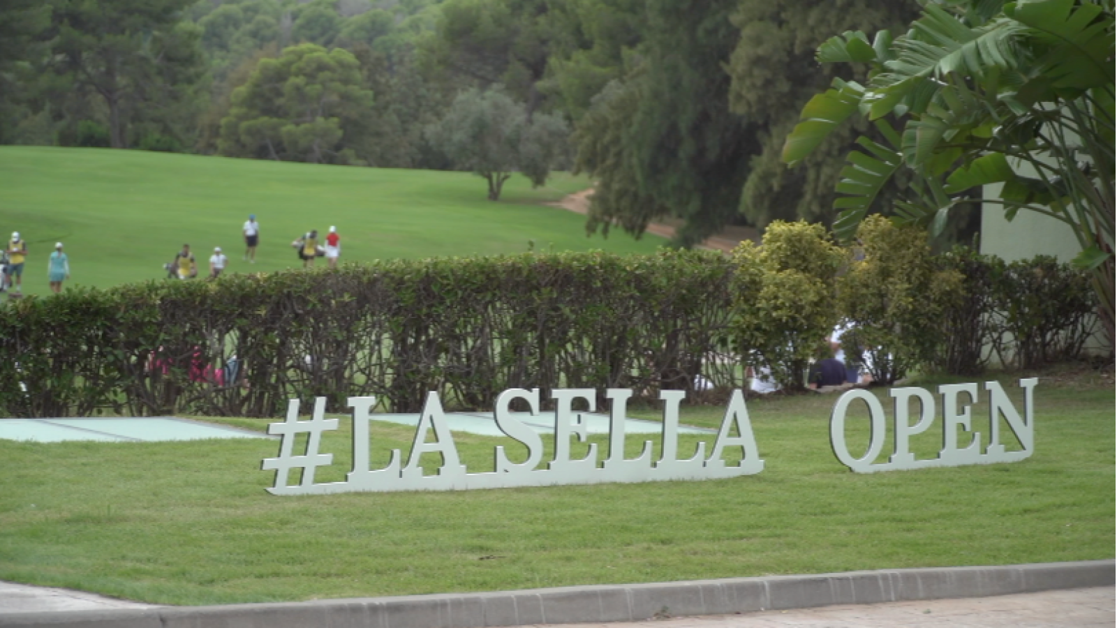 Un campo de golf con césped verde y árboles al fondo, con personas jugando en la distancia. Un letrero promocional en primer plano anuncia un evento deportivo.