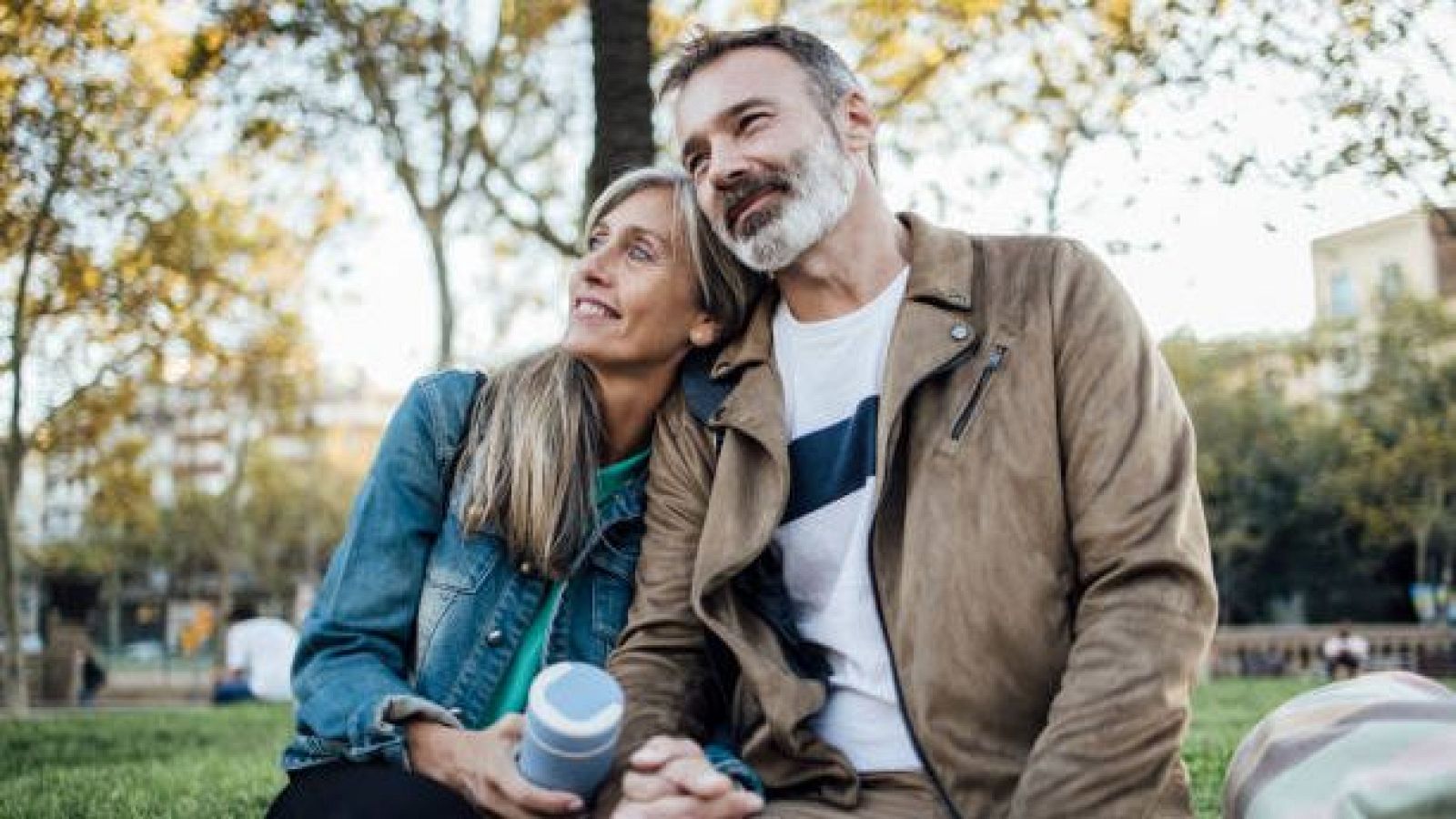 Una pareja madura disfruta de un momento romántico en un parque, con la mujer apoyando su cabeza en el hombro del hombre. Ella lleva una chaqueta vaquera y él una chaqueta marrón, ambos mirando hacia arriba con una sonrisa.