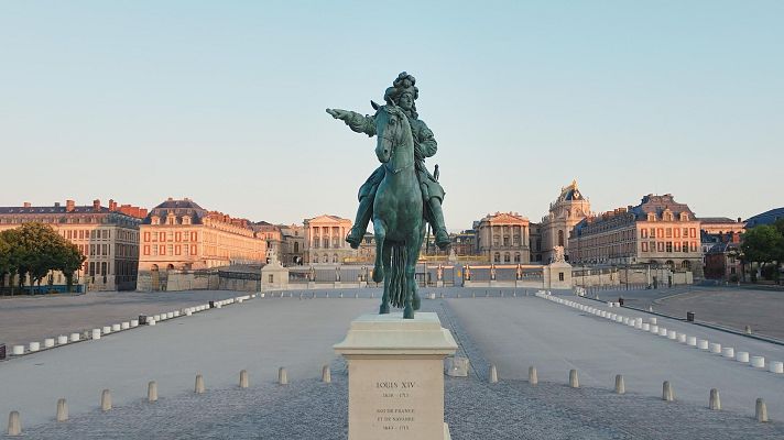 Una estatua ecuestre de un rey, con vestimenta detallada y una pose imponente, se alza en una plaza pavimentada. La base de la estatua muestra una inscripción con fechas y títulos. Al fondo, se vislumbran edificios clásicos.