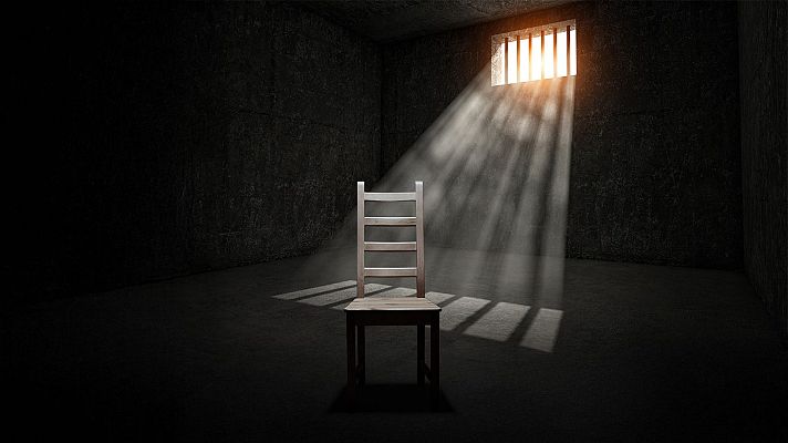 Una celda oscura con paredes de piedra y una ventana enrejada, iluminada por un haz de luz que proyecta sombras en el suelo. En el centro, una silla de madera simple se encuentra parcialmente iluminada, evocando una sensación de aislamiento.