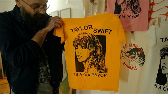 Un hombre con barba y gafas muestra una camiseta amarilla con la imagen de una persona y la frase \"TAYLOR SWIFT IS A CIA PSYOP\". En el fondo, se aprecian más camisetas con mensajes similares y diseños variados.