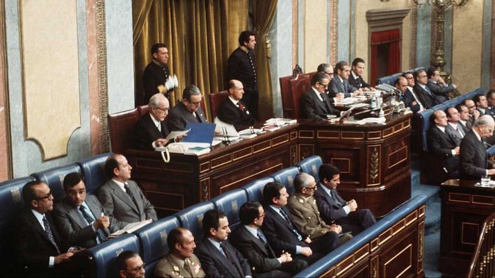 En una sala de estilo clásico, con asientos azules en hemiciclo, se observa un debate parlamentario con hombres vestidos formalmente, algunos con gafas y tomando notas, presumiblemente en 1976.