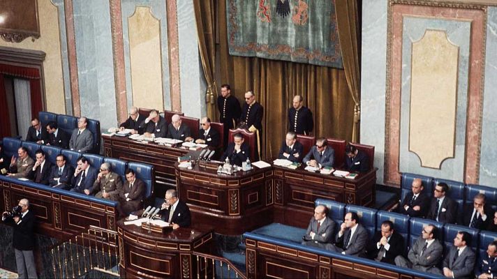 Un salón de plenos con asientos azules y cortinas doradas es el escenario de un debate político. Diputados y personal de apoyo están presentes, con un fotógrafo capturando la escena.