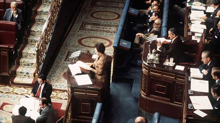 Imagen de un salón de plenos con escaños de madera y alfombras con motivos geométricos. Se observa a un hombre escribiendo en una mesa y a una mujer leyendo un documento en la tribuna, ambos en un ambiente formal.
