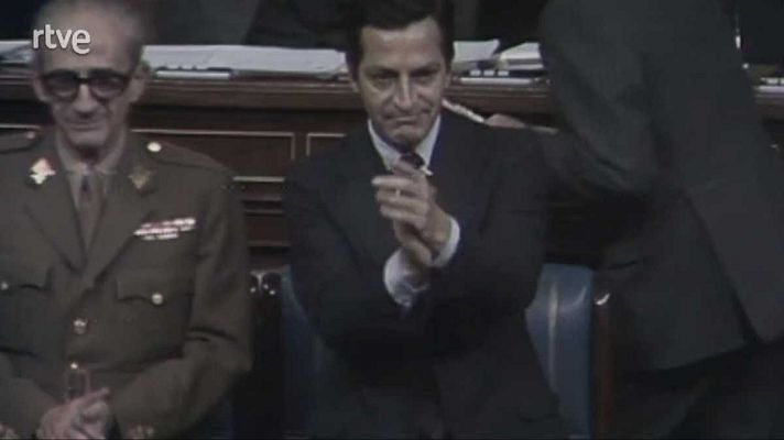 En una sala, el general Gutiérrez Mellado, uniformado, está sentado mientras Adolfo Suárez, con traje, aplaude a su lado.