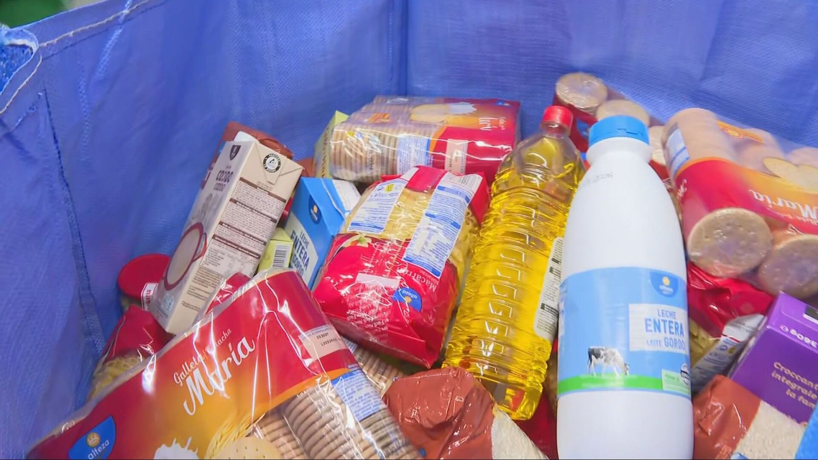 La imagen presenta una bolsa azul llena de alimentos, incluyendo galletas, aceite, leche y pasta, posiblemente recolectados para una campaña de donación.