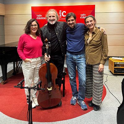 Cuatro personas sonríen en un estudio de radio, con un piano de cola y un panel rojo de fondo. Un hombre con un violonchelo, posiblemente José el Marqués, y una mujer con gafas destacan en la imagen.