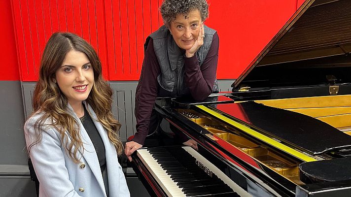Dos mujeres, ambas identificadas como M.ª Ángeles Ayala, posan junto a un piano de cola negro con detalles dorados. Una está sentada frente al piano, vestida con ropa oscura y una chaqueta clara, mientras que la otra está de pie, apoyada en el piano.
