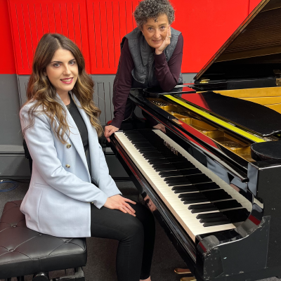 Dos mujeres, ambas identificadas como M.ª Ángeles Ayala, posan junto a un piano de cola negro con detalles dorados. Una está sentada frente al piano, vestida con ropa oscura y una chaqueta clara, mientras que la otra está de pie, apoyada en el piano.