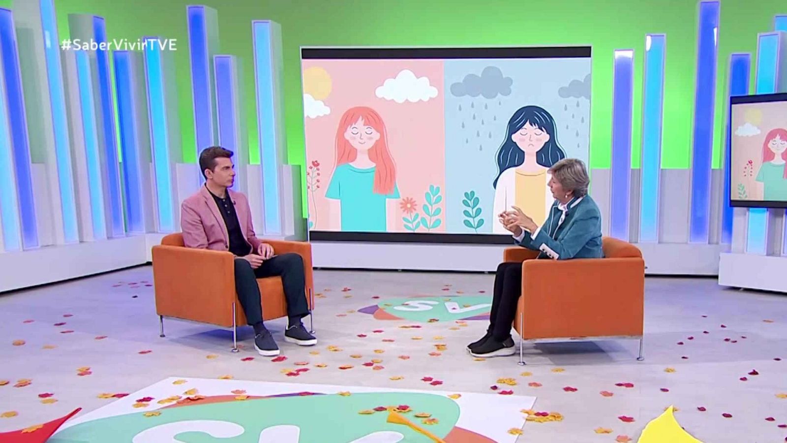 En un plató televisivo, un hombre con chaqueta rosa y una mujer con chaqueta azul están sentados en sillones, con pantallas que muestran ilustraciones de diferentes climas. El suelo está decorado con confeti.