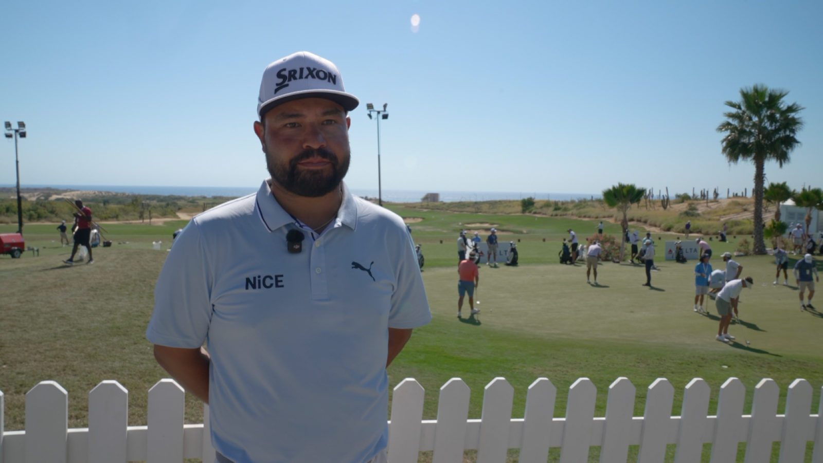 Un hombre con gorra y camisa con logos, posa en un campo de golf con palmeras y cactus, con el mar visible al fondo, y una valla blanca en primer plano.