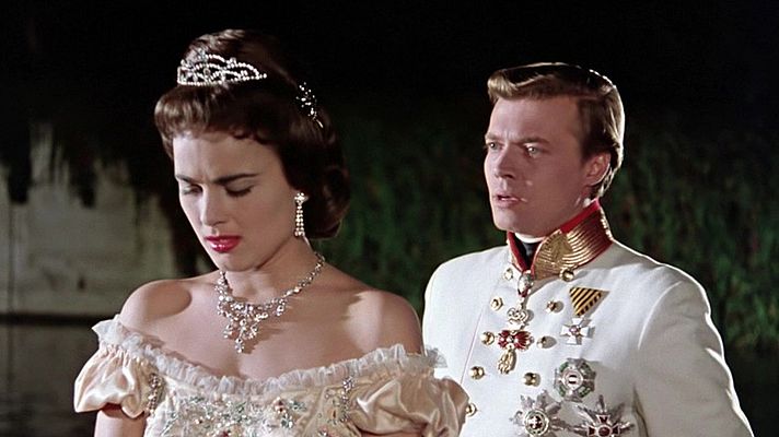 Sissi, con un vestido elegante y joyas, comparte escena con un hombre en uniforme militar. Ambos parecen estar en un entorno exterior oscuro, posiblemente un jardín.