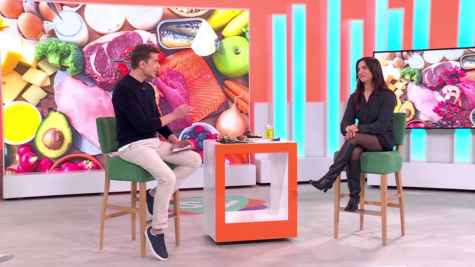 En el plató de un programa de televisión, dos personas están sentadas frente a una mesa con alimentos. Un hombre con jersey oscuro gesticula, mientras que una mujer con vestido oscuro y botas negras está sentada con las piernas cruzadas.