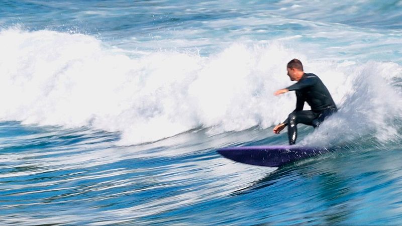 Un surfista con traje de neopreno negro se desliza sobre una tabla morada en el mar, enfrentándose a olas grandes y espumosas. El agua es azul intenso y se puede leer un texto en la imagen.