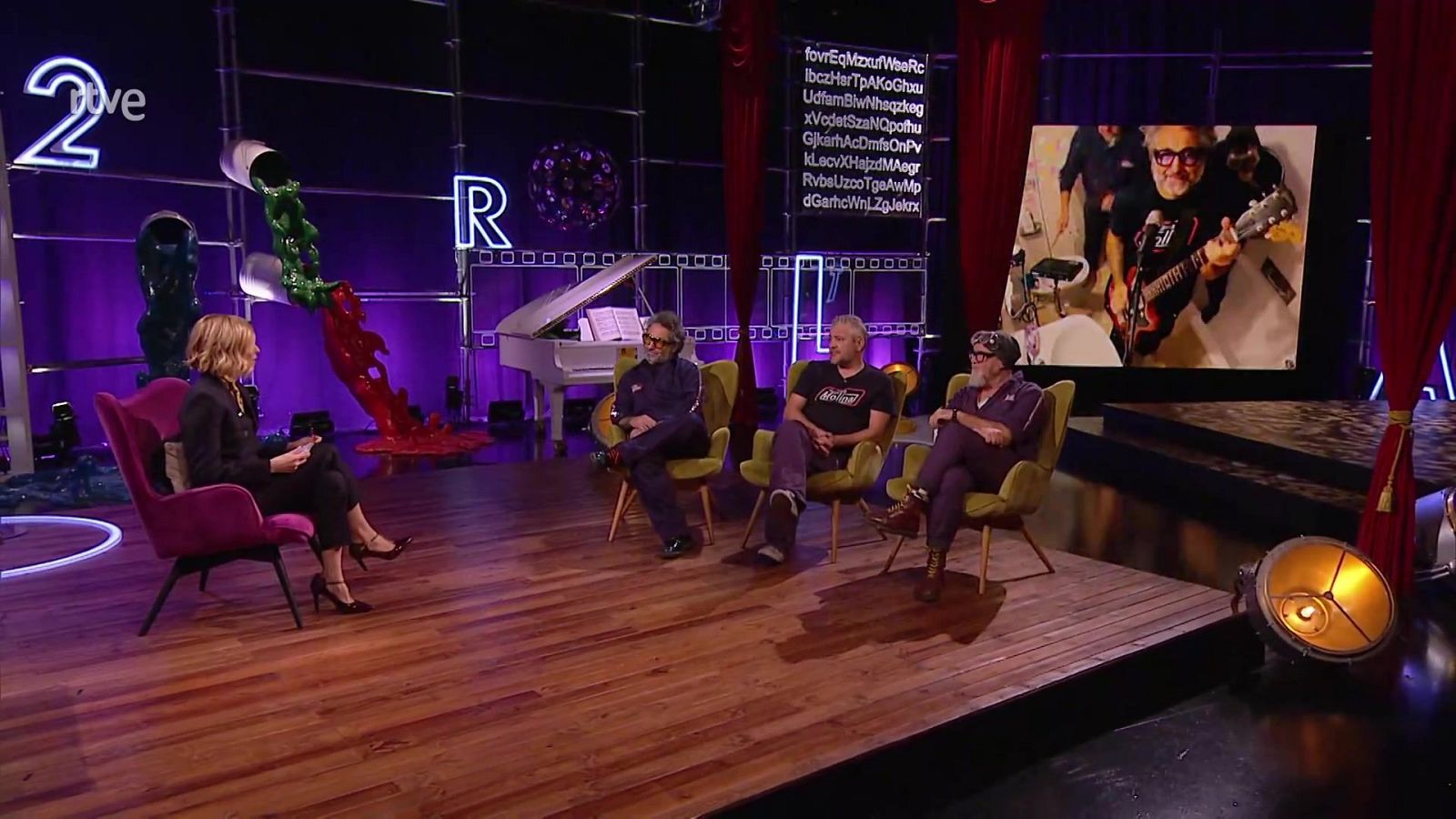 En un estudio televisivo, una presentadora en un sillón fucsia conversa con tres hombres sentados. El decorado incluye elementos artísticos y una pantalla que muestra a un músico tocando.