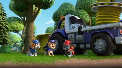 Tres cachorros con cascos y uniformes de construcción se encuentran frente a un camión azul y blanco con un carrete de cable. El entorno es un área boscosa con árboles y hierba.