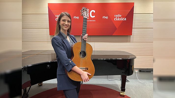 Una mujer sonriente, identificada como Raquel del Pino, posa con una guitarra clásica, vestida con una chaqueta azul marino y una blusa estampada, con un piano de cola y el logotipo de Radio Clásica RTVE al fondo.