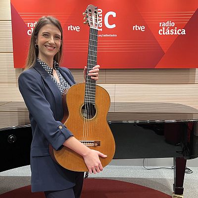 Una mujer sonriente, identificada como Raquel del Pino, posa con una guitarra clásica, vestida con una chaqueta azul marino y una blusa estampada, con un piano de cola y el logotipo de Radio Clásica RTVE al fondo.