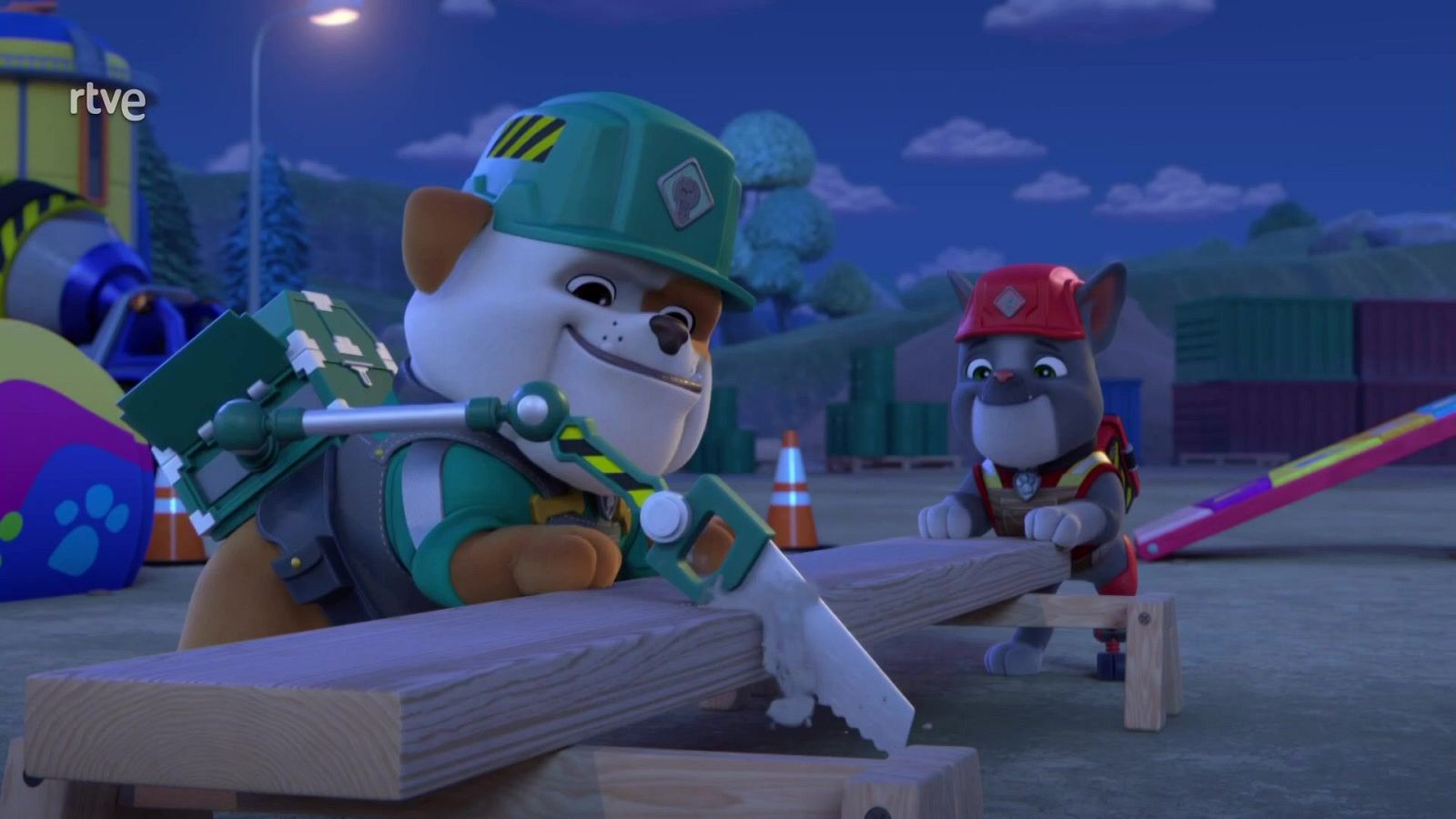 Rubble, un cachorro con casco verde, corta madera con una sierra mientras otro cachorro con casco rojo sujeta la tabla en un entorno de construcción con conos y contenedores.