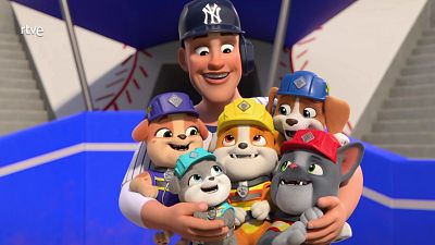 Un hombre con gorra de béisbol y uniforme abraza a cachorros con cascos de colores, posiblemente en un estadio. Uno de los cachorros es Rubble.