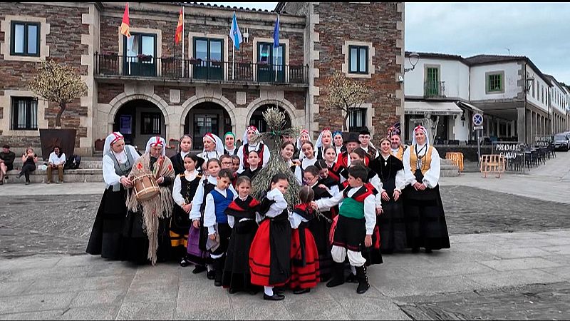 Un grupo de personas vestidas con trajes tradicionales se congrega en una plaza empedrada. Algunos llevan instrumentos musicales, y al fondo se observa un edificio con banderas y un letrero.