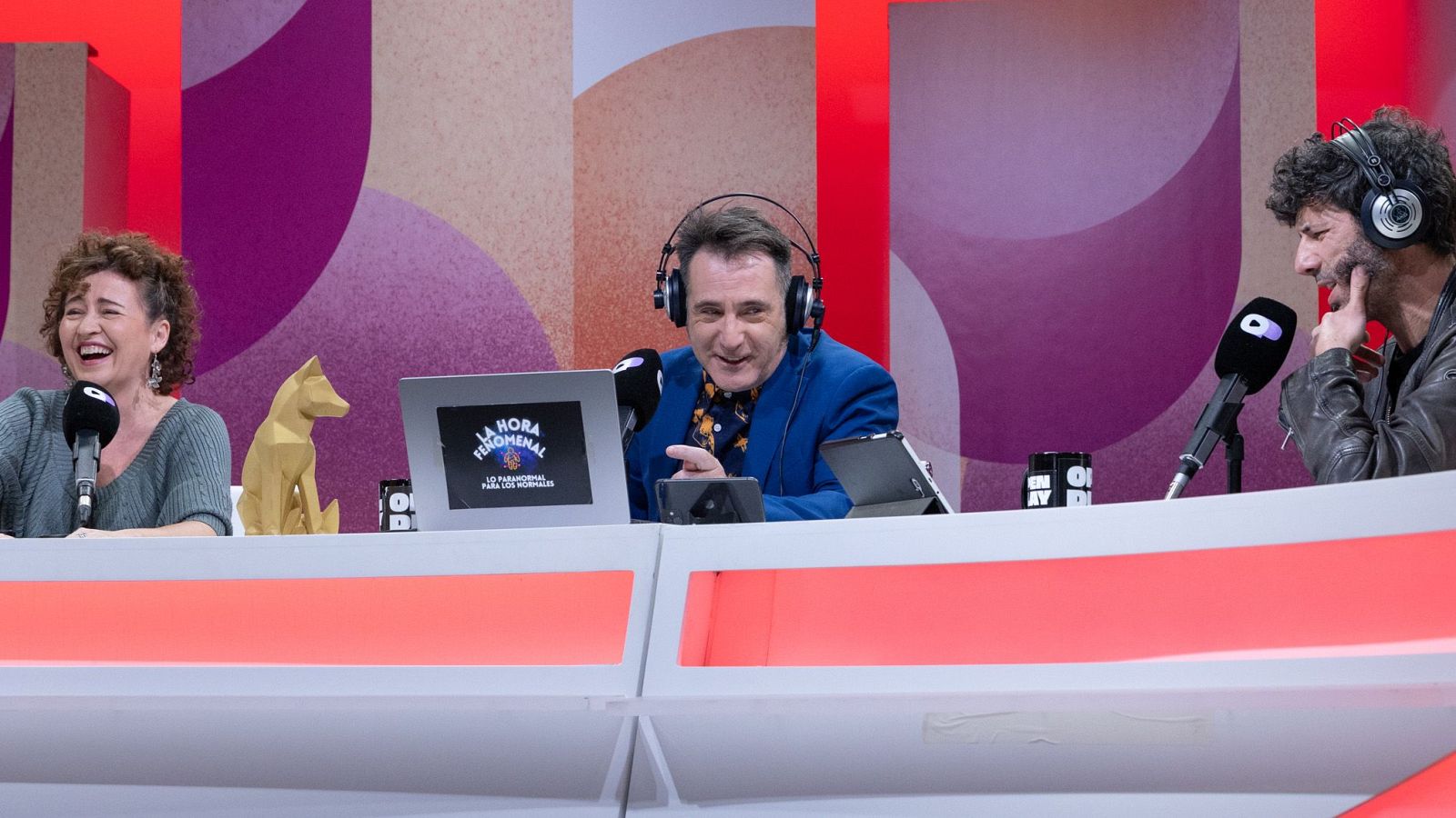 Tres individuos se encuentran en un estudio, posiblemente grabando un podcast. Uno sonríe y señala, otro ríe y el tercero parece reflexionar, con micrófonos y ordenadores frente a ellos, y una pared colorida de fondo.