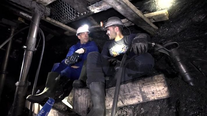 En una mina de carbón, dos hombres con cascos y linternas dialogan sentados. Uno viste mono azul y botas verdes, el otro ropa oscura y botas negras, en un ambiente polvoriento con elementos de soporte y una perforadora.