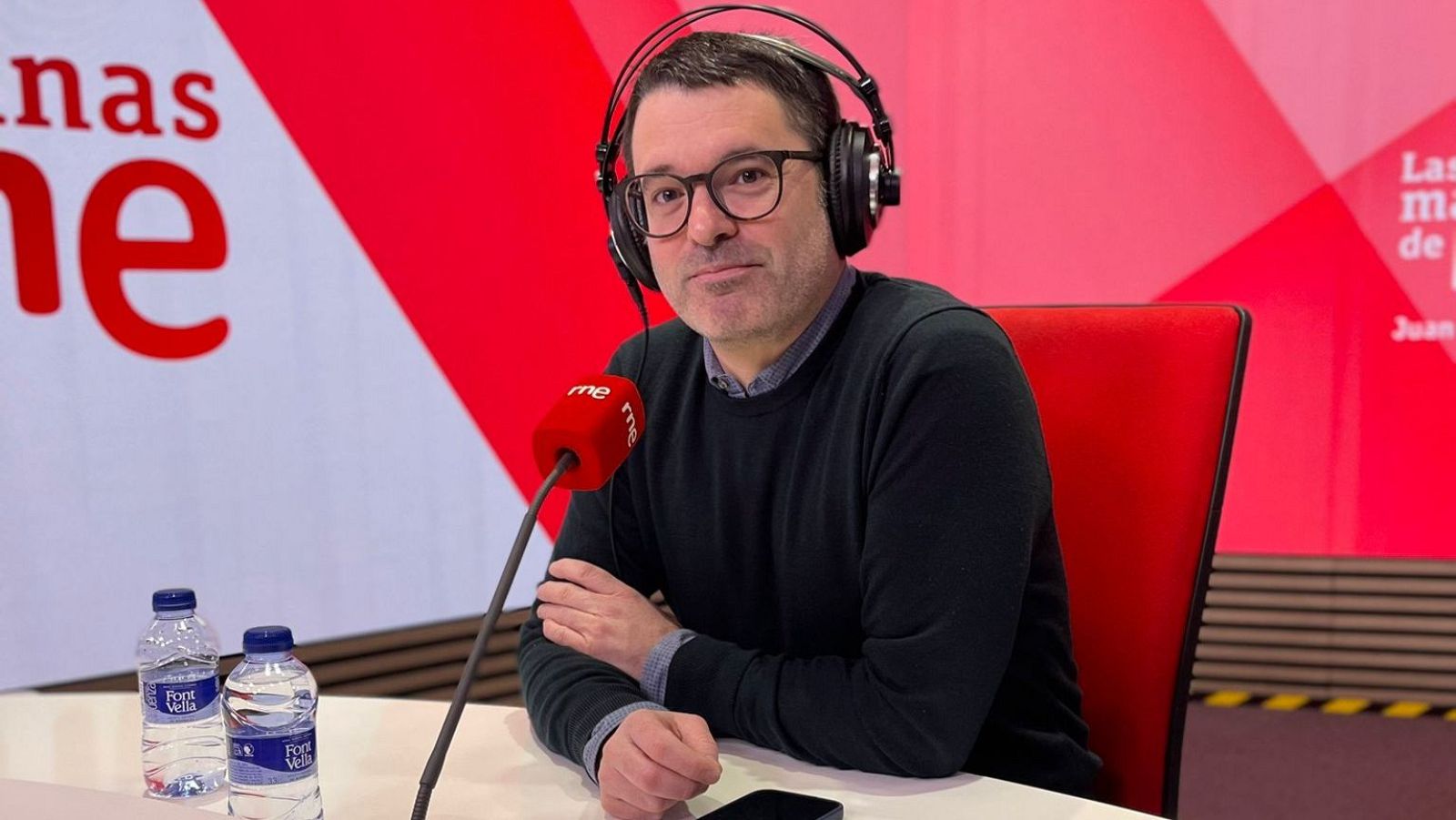 Ramón González Férriz, con auriculares y jersey oscuro, está sentado frente a un micrófono en un estudio de radio, mirando a la cámara con una expresión tranquila. Se observan botellas de agua y el logo del programa al fondo.