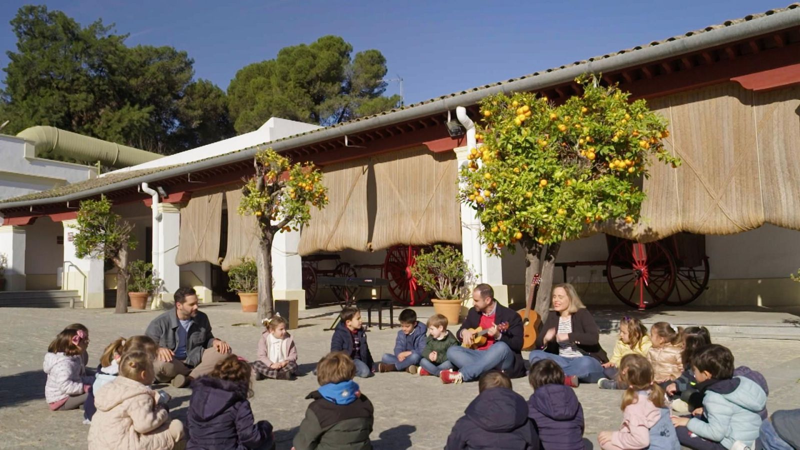 En un patio, un grupo de niños escucha atentamente a un hombre que toca un ukelele, con una mujer adulta presente. El escenario se completa con un edificio, árboles y carros antiguos.