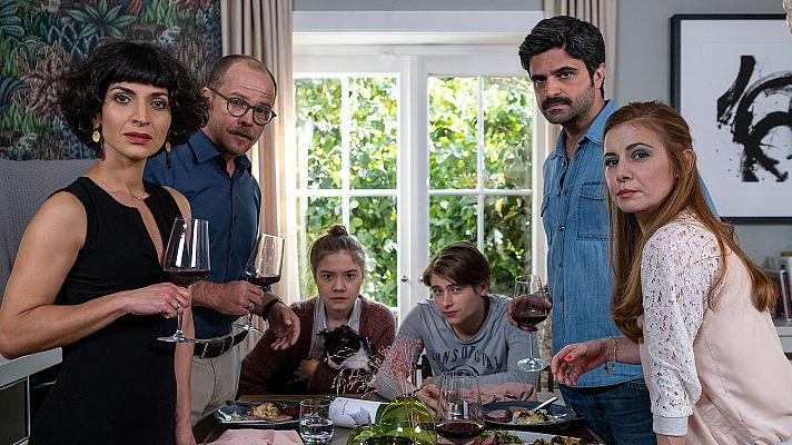 Una mesa de comedor es el centro de la imagen, con ocho personas reunidas. Se observan detalles como copas de vino, platos de comida y una mujer con vestido negro, junto a una mujer pelirroja. El ambiente sugiere una escena de una película.