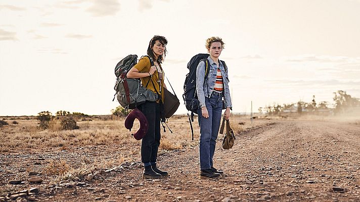 En un entorno desértico, dos mujeres jóvenes, una con mochila verde y otra con mochila azul, se encuentran en un camino de tierra. Una lleva camiseta mostaza y la otra, una camiseta a rayas, con el cielo nublado y polvo en el ambiente.